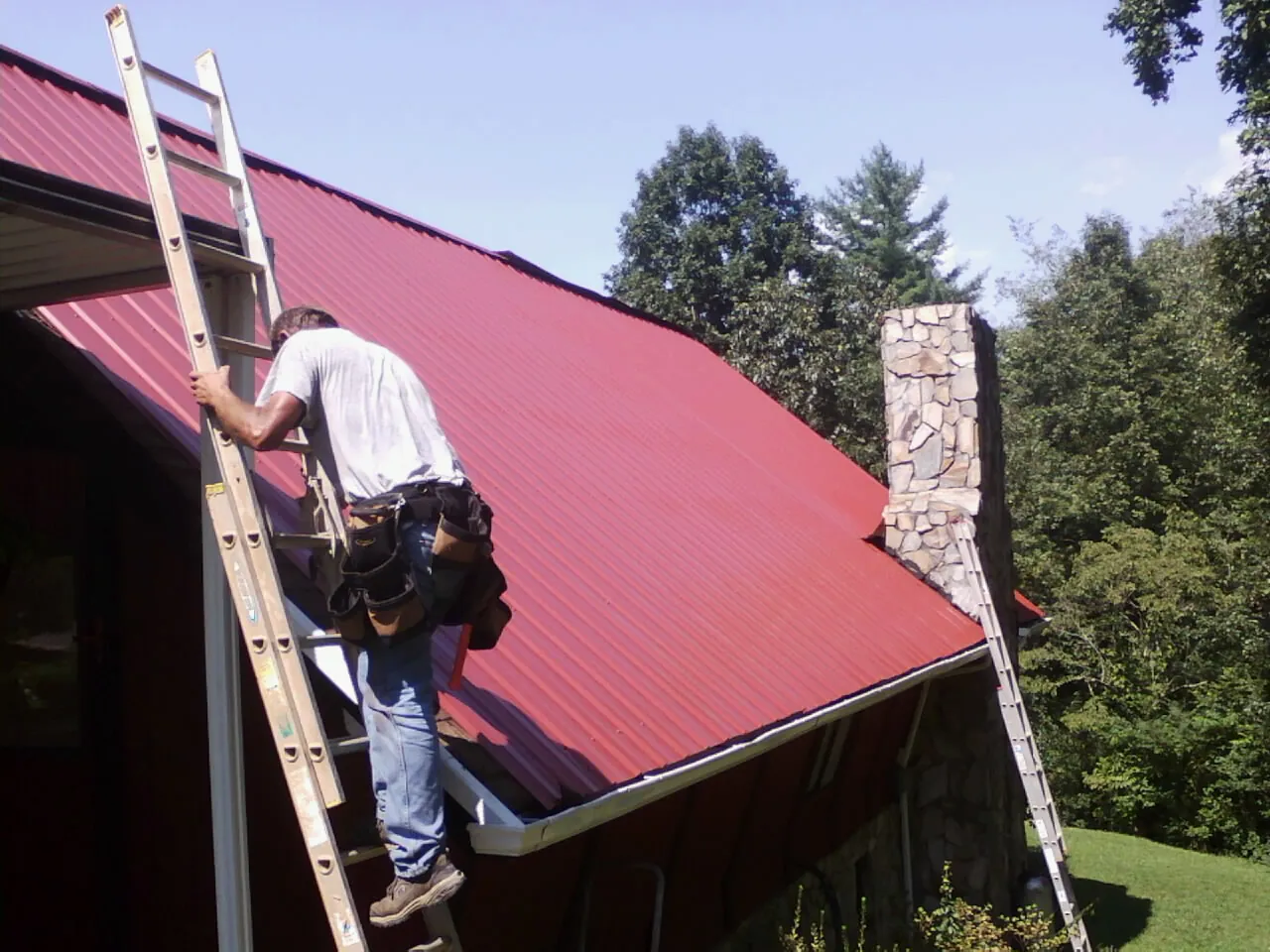 Roofer on a ladder beside a red metal roof and stone chimney — Soffit & Fascia Repair by Roofing Brothers in Diamondhead